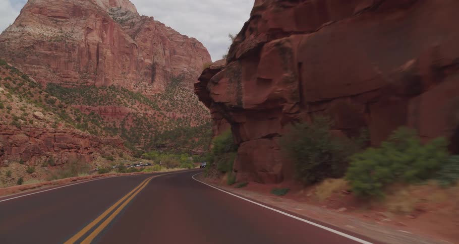 A vehicle view drives along a curving two lane road through a deep mountain canyon with towering red rock cliffs slopes covered in green brush and oncoming cars under an overcast sky
