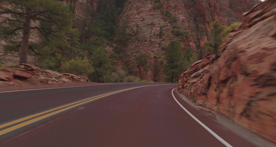 A vehicle view drives along a red asphalt road through a mountain canyon passing oncoming cars and surrounded by towering red and tan rock cliffs and scattered green trees under an overcast sky