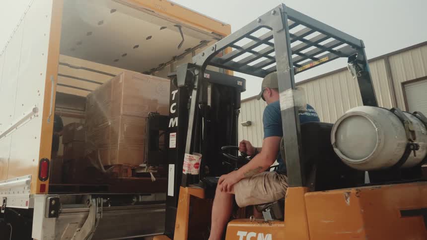 A man is driving a forklift and lifts a large box out of the back of a semi truck