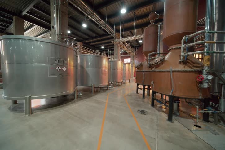 A view down an aisle in a large industrial facility multiple rows of large silver stainless steel tanks occupy the left several tall brown copper stills and columns fill the right the floor has yellow striping pipes run overhead