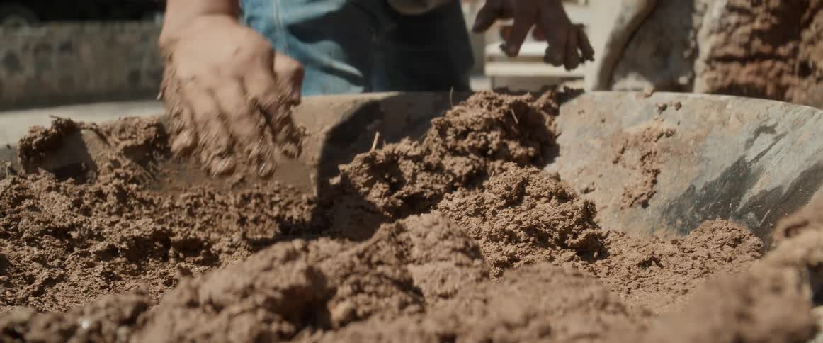 A closeup view of two hands mixing a moist brown earthen material the material is contained within a metal wheelbarrow tray the lower body of a person wearing jeans stands behind the tray the background shows a paved ground and a vehicle