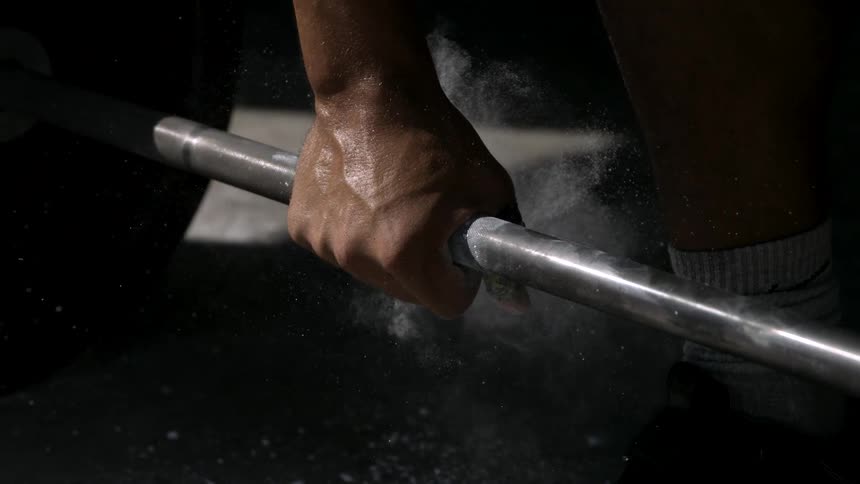 A hand moving to grip a silver barbell white chalk dust flies up from the hand and bar the barbell section is covered in chalk residue a persons lower leg and socks are visible on the right the area is dark with focused lighting