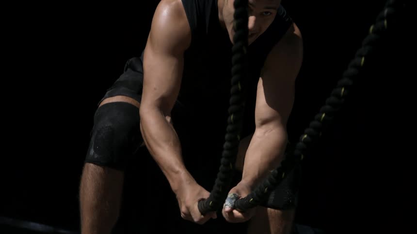 A strong man wearing a black tank top and knee pads vigorously slams heavy battle ropes on the ground as part of an exercise routine in a dimly lit gym