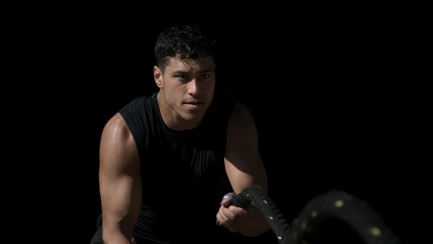 The video is a medium closeup of a muscular man doing battle rope exercises looking determinedly forward against a black background he is wearing a dark sleeveless workout shirt and the lighting highlights his arms and shoulders