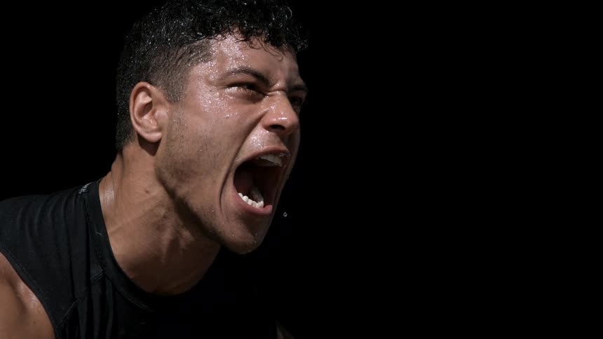 A sweaty man shouts with mouth wide open neck veins standing out as his face contorts in intense exertion against a black background