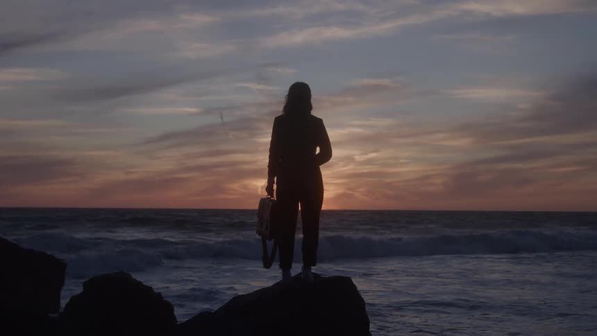 The video shows a woman standing in silhouette on a rocky surface next to the ocean at sunset or sunrise she is facing away from the camera and holding a briefcase in one hand and possibly a circular helmet or object in the other