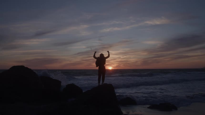 Woman standing on a rock along the coast stretching her arms in the air as waves roll into shore and the sun glows on the horizon