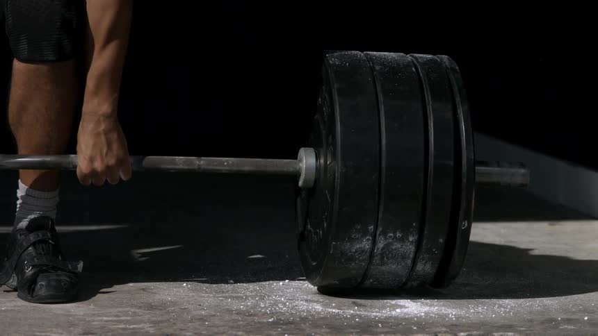 A persons lower body and a loaded barbell on the floor the person bends down to grip the bar the heavy barbell is lifted off the ground white chalk dust sprinkles down onto the concrete floor the area is dark with a focused light source