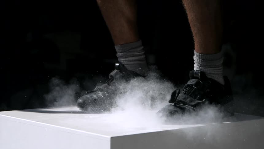 A man wearing black sneakers jumps onto a square platform as white powdery dust rises off the surface