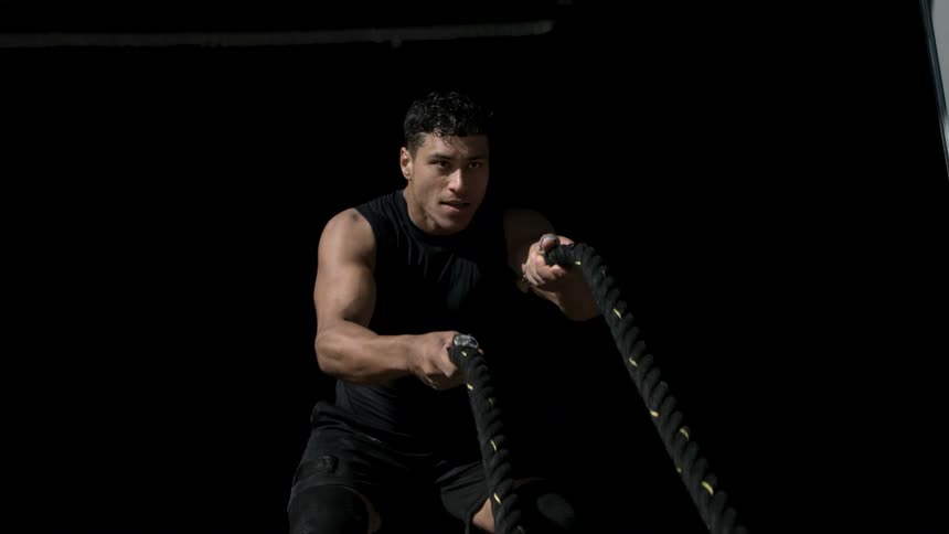 A man in a sleeveless black top drives battle ropes in a dark space face intent and muscles taut