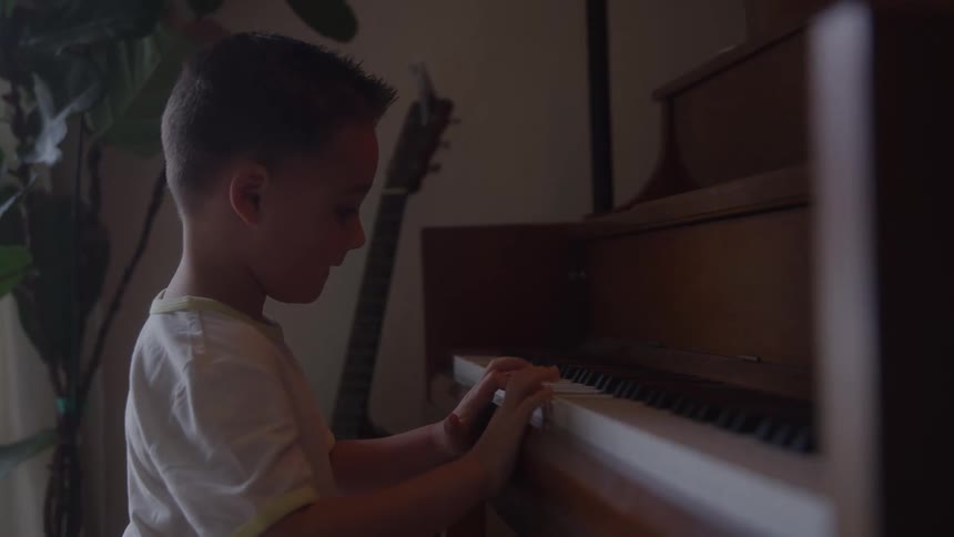 A young boy wearing a white tshirt plays a dark upright piano with concentration in a dimly lit room where a guitar is visible in the background
