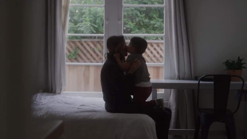 A mother and son share a tender kiss while sitting in a softly lit bedroom