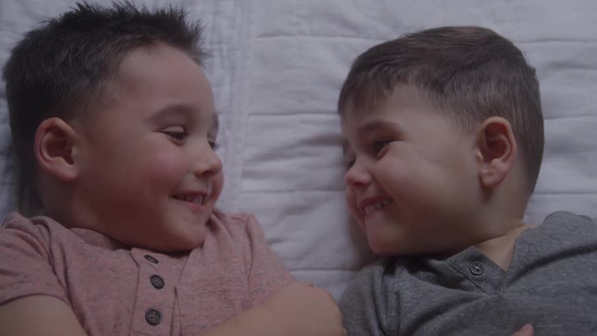 Two young boys lie facing each other on a white quilted blanket the boy on the left wears a pink shirt the boy on the right wears a gray shirt both boys look at each other and begin smiling widely