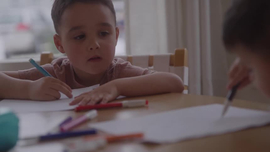 A boy sitting with a colored pencil in his hand watching another boy drawing on a sheet of paper