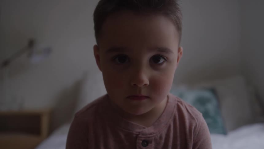 A child wearing a pink shirt sits on a bed inside a room at midday