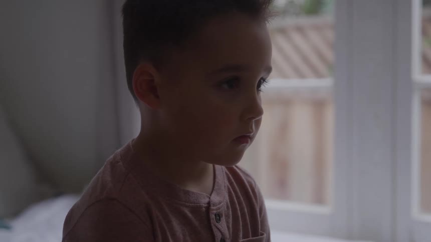 A child sits quietly on a bed in a bedroom at midday their expression marked by sadness as soft daylight filters through the window