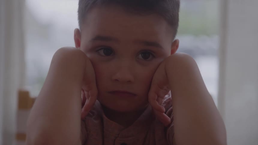 A close view shows a young boy with a serious expression and a pink shirt resting his head in his hands against a light colored background
