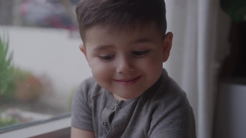 The boy is looking down and smiling by the window