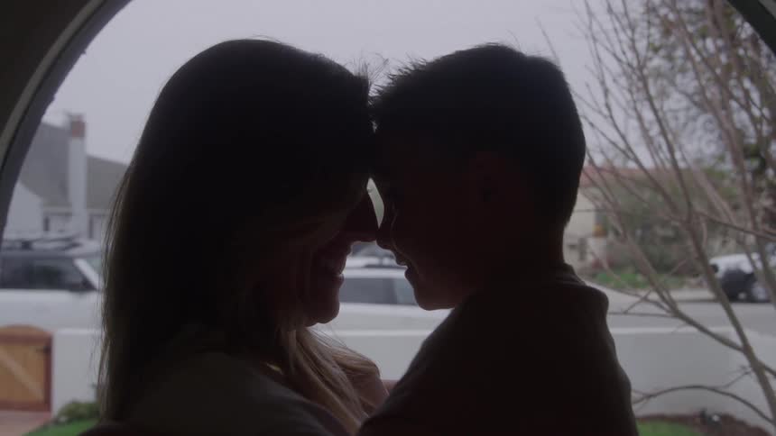 A silhouette close view shows a man and woman standing close and smiling at each other their foreheads touching near a doorway in a soft bright outdoor light with a suburban street visible behind them