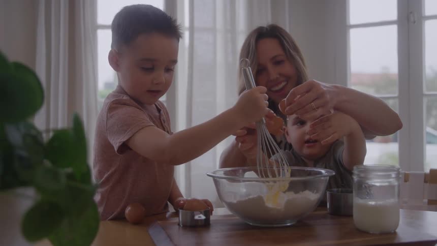 A family gathered around a table baking a cake a mother with a small child adding eggs to the batter and a boy mixing the batter with a whisk