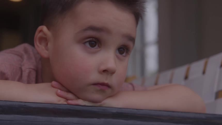A close view shows a young boy wearing a pink shirt resting his chin on his crossed arms on a dark window sill with an attentive look on his face