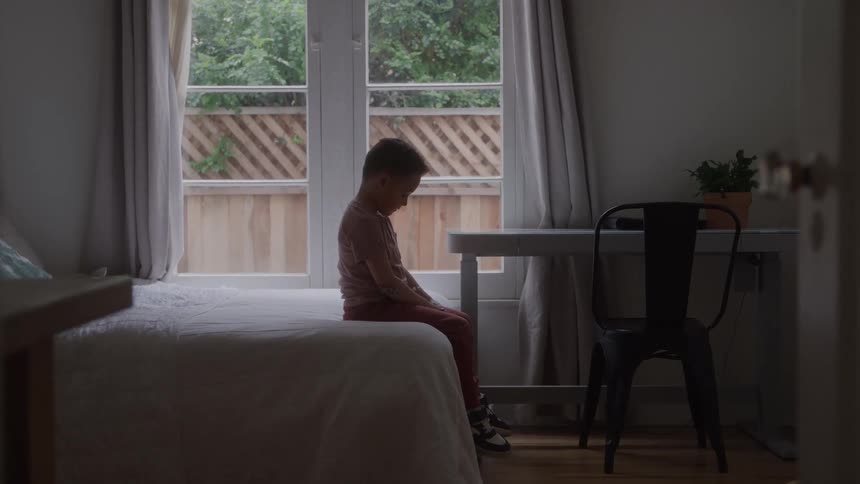 A young boy wearing a pink shirt and red pants sits alone on the edge of a bed looking down sadly with his hands clasped in a dim bedroom with glass doors leading outside