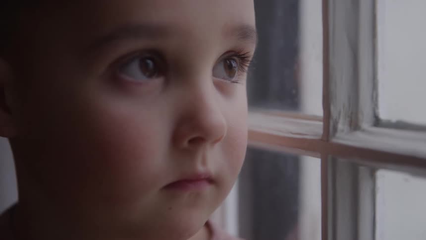 A closeup of a young boy gazing out a window inside his home
