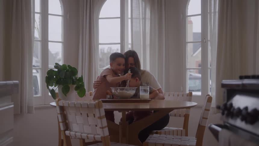 Mother hugs her two sons while the eldest one mixes the contents of a bowl during the daytime