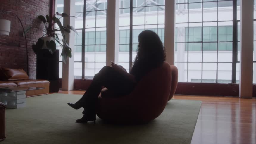 A woman sits silhouetted in a large curved chair in a spacious loft the woman is interacting with a smartphone a tall wall of multipaned windows occupies the background the room contains furniture