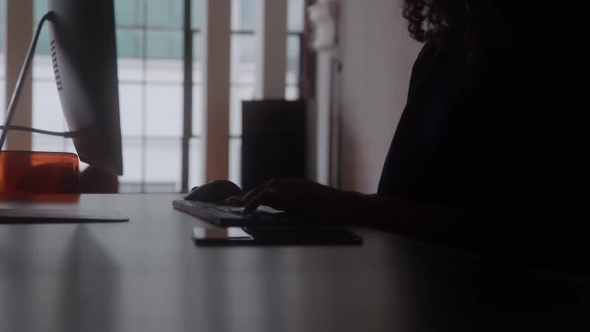 A woman sits at a desk at home while typing on a computer keyboard