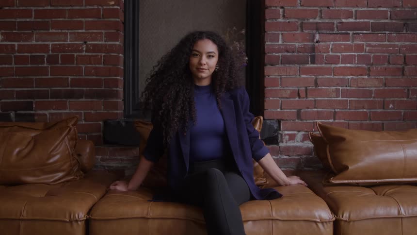 A woman sits on a leather sofa looks at the camera a brick wall forms the background a dark window opening is centered behind her and she wears a dark blazer