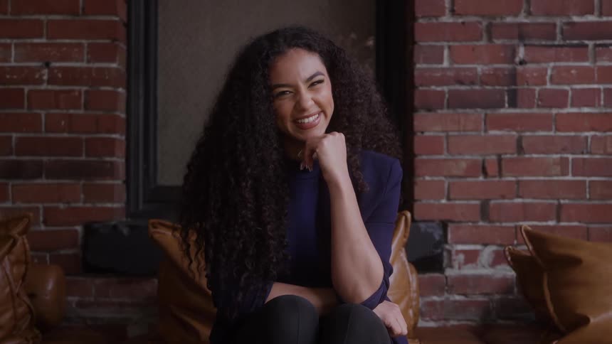 A woman with long curly hair smiles warmly as she sits on a leather couch against an exposed brick wall