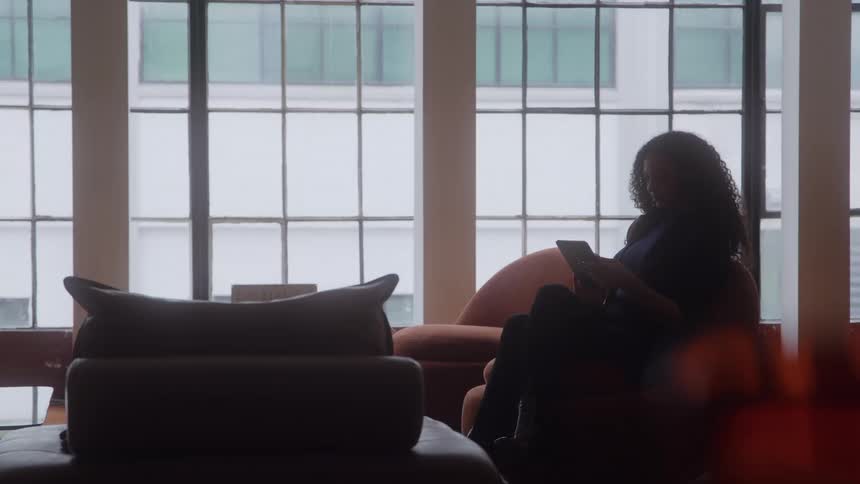 A woman sits near large windows using a tablet in a softly lit office lobby