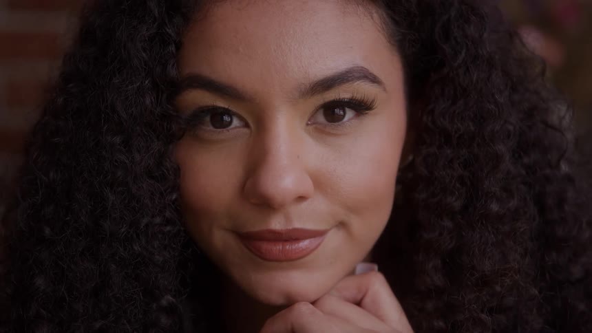 The video is a tight closeup of a young woman with long dark curly hair she initially looks offcamera with a contemplative expression before turning to look directly at the camera with a slight smile