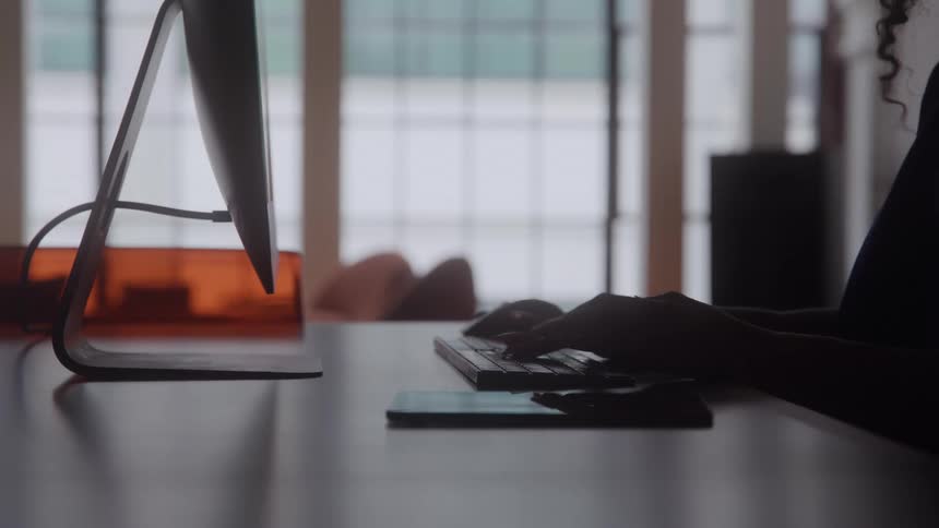 The video shows a closeup of a persons hands and arms silhouetted while they are typing on a keyboard at a desk the scene is dimly lit indoors with the person facing a bright gridpaned window in the background