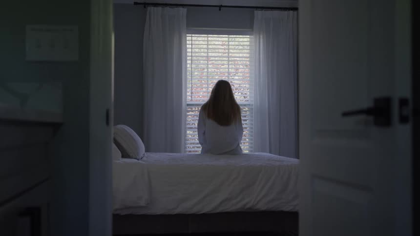 A woman sits alone on a bed facing a window in a quiet dimly lit bedroom