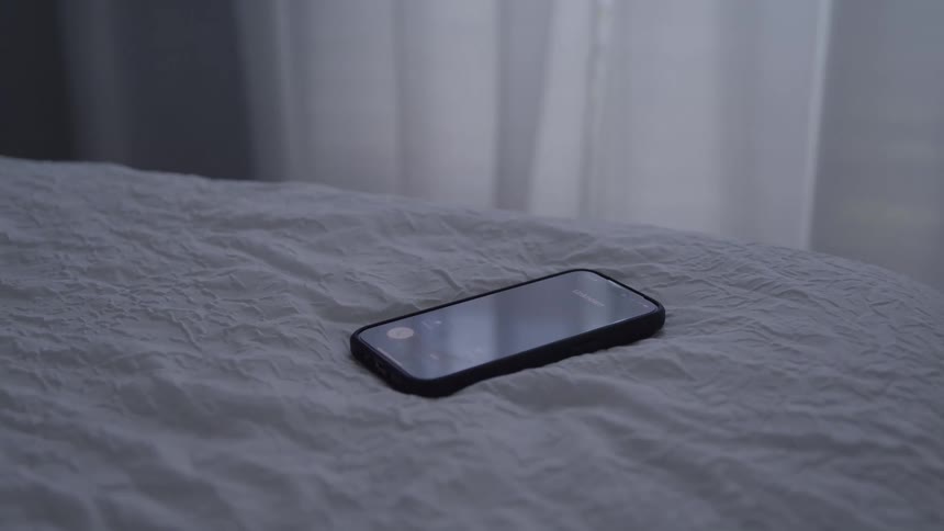 A hand picks up a smartphone from a bed in a softly lit bedroom