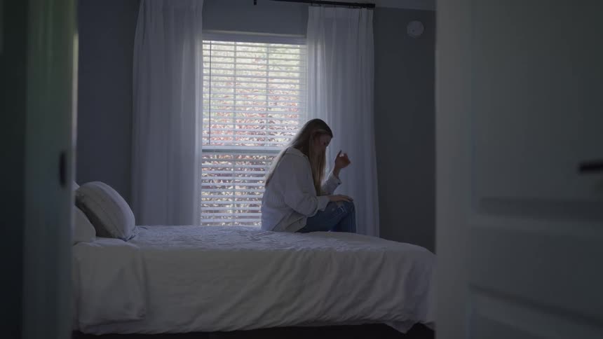 A sad woman wearing a white shirt and jeans sits alone on a white bed looking down with her arms hugged to her knees in a dim room with bright light coming through the window blinds