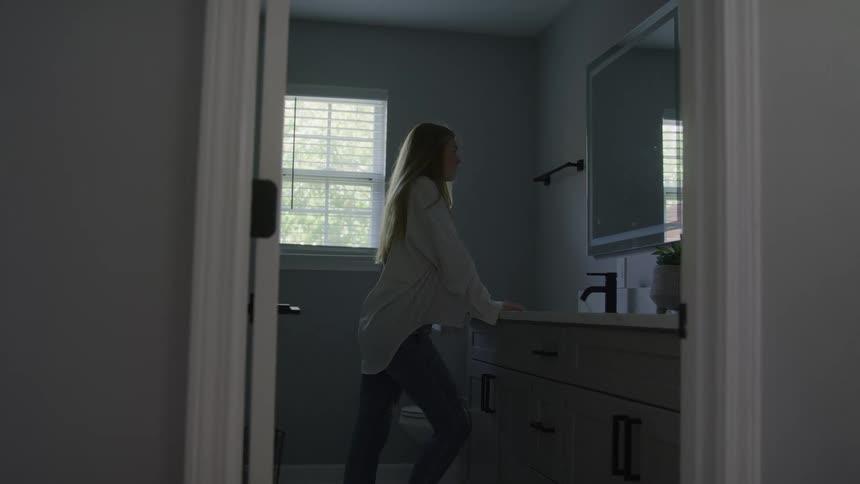 Blonde woman standing with her hands on the bathroom counter while watching her reflection drops her head forward