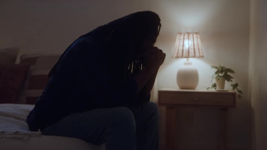 Man sits on the edge of the bed with hands clasped before a lit lamp