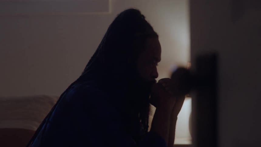 Man with braided hair sitting on bed with hands near face in room lit by table lamp