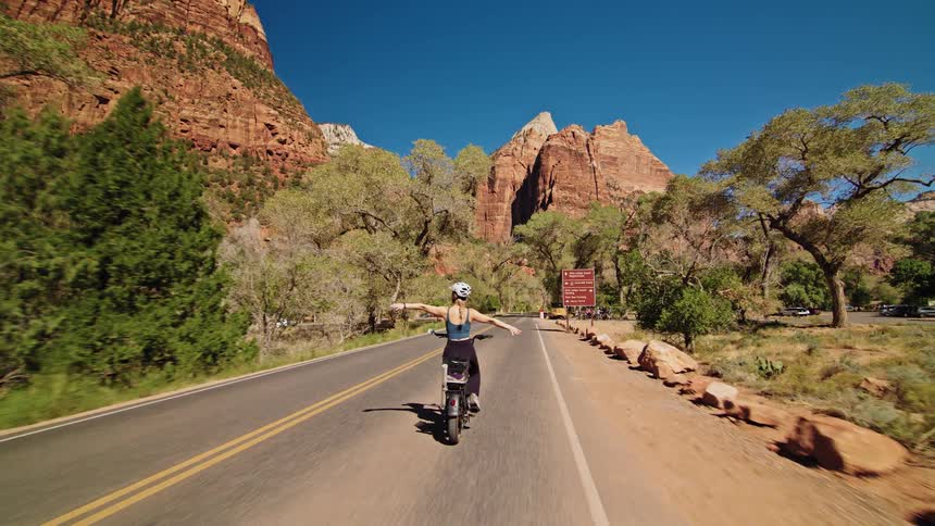 A joyful woman wearing a helmet rides an electric bike down a park road spreading her arms wide in front of massive red sandstone cliffs and green trees under a clear blue sky