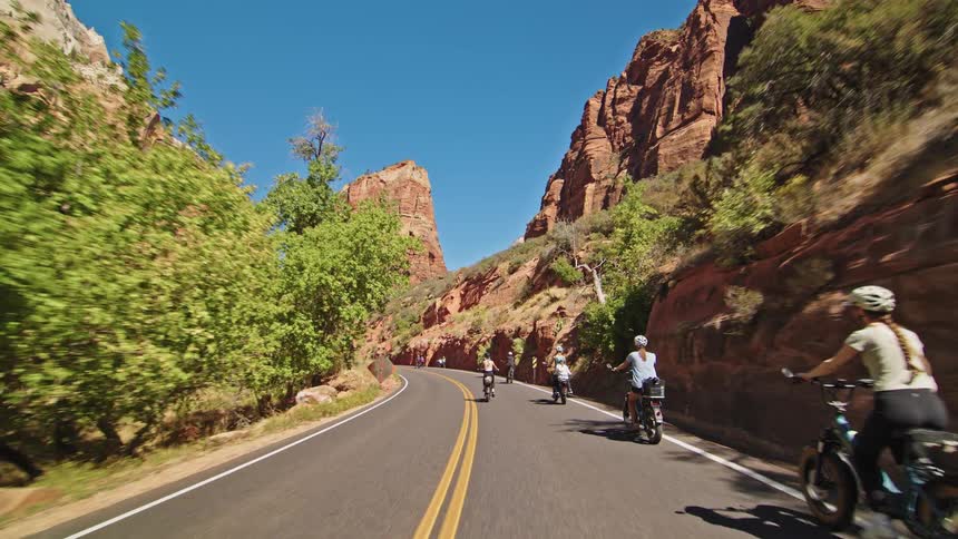 A group of cyclists including a mother with a child in a rear seat enjoy a sunny ride on a winding road through a magnificent red rock canyon