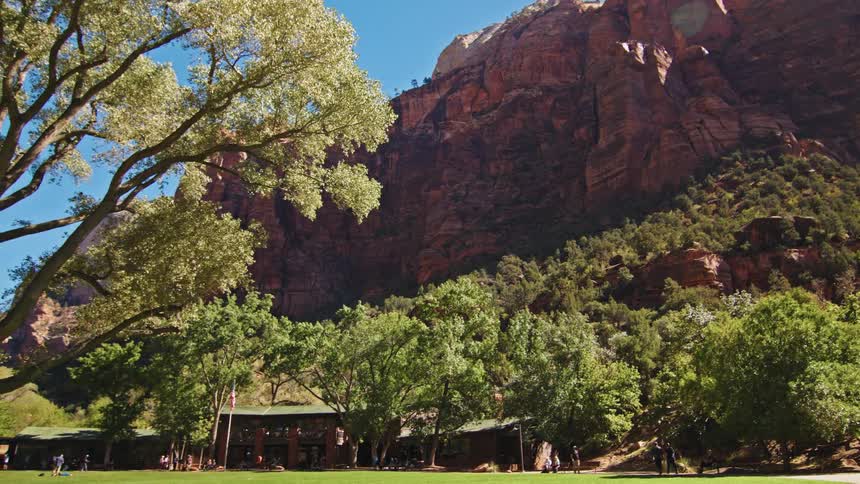 A sprawling green lawn and lodge are nestled at the base of a magnificent sunlit red rock canyon