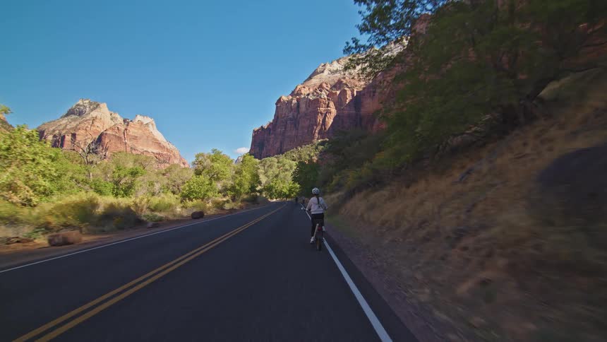 A woman wearing a white hoodie and a helmet rides a bicycle down a paved road surrounded by immense red sandstone cliffs and vibrant green foliage on a sunny day
