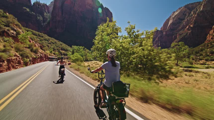 A woman rides a bike along the side of a highway with other helmeted cyclists all making way for a passing bus as rocky terrain lines the road