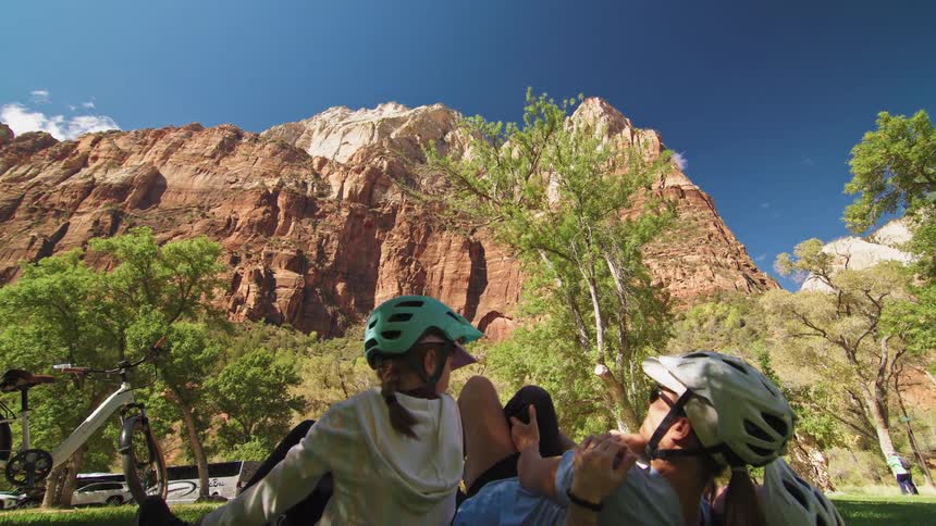 Two women and a young girl sit on a grassy lawn in a public park all wearing helmets after cycling with a scenic mountain view stretching behind them under the bright midday sun