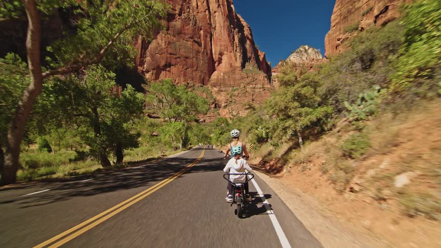 A young woman rides a bike along the side of a freeway on a sunny day heading toward the grand canyon with a child seated behind her during the journey