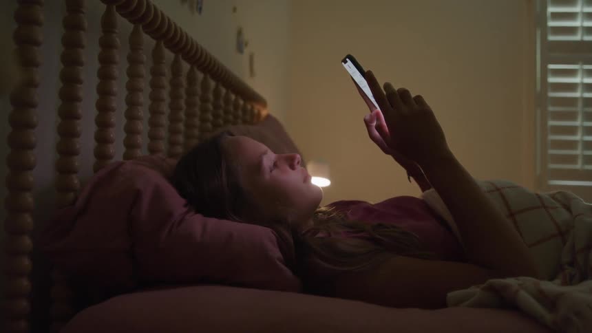 Girl lying in bed holding her phone in a dimly lit room