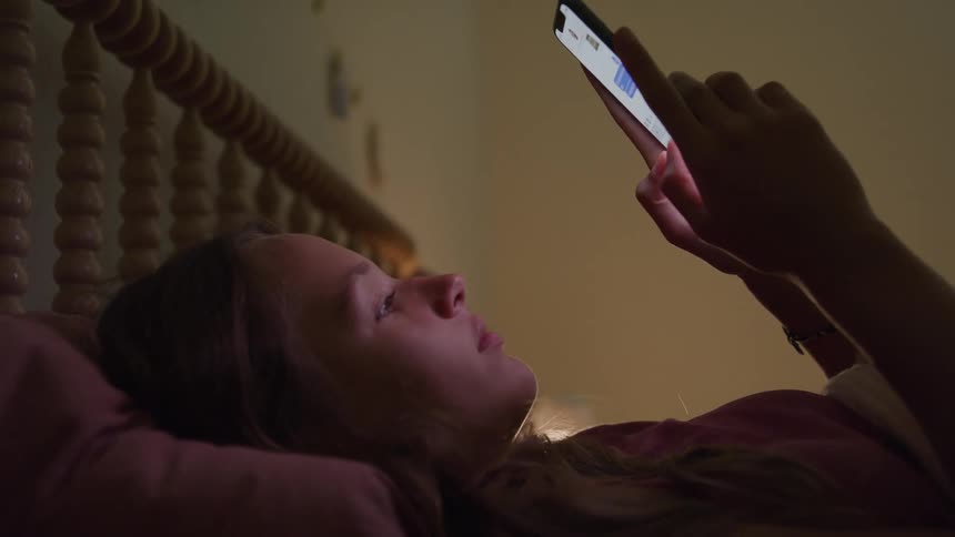 A young girl is laying in bed texting on a smart phone